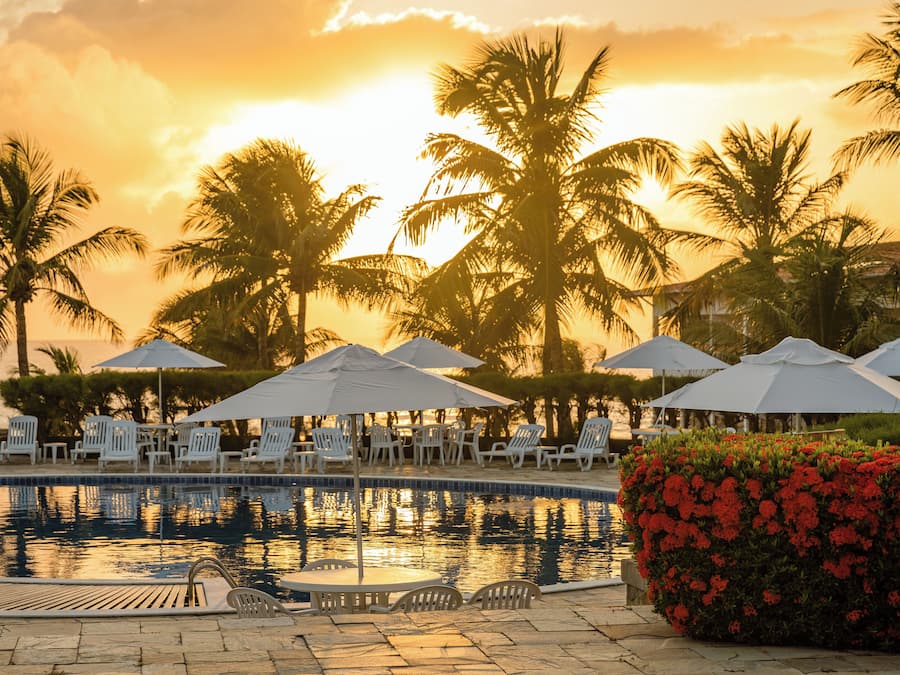 Un espace piscine agrémenté de chaises longues et de parasols, entouré de palmiers et offrant une vue imprenable sur le coucher de soleil.