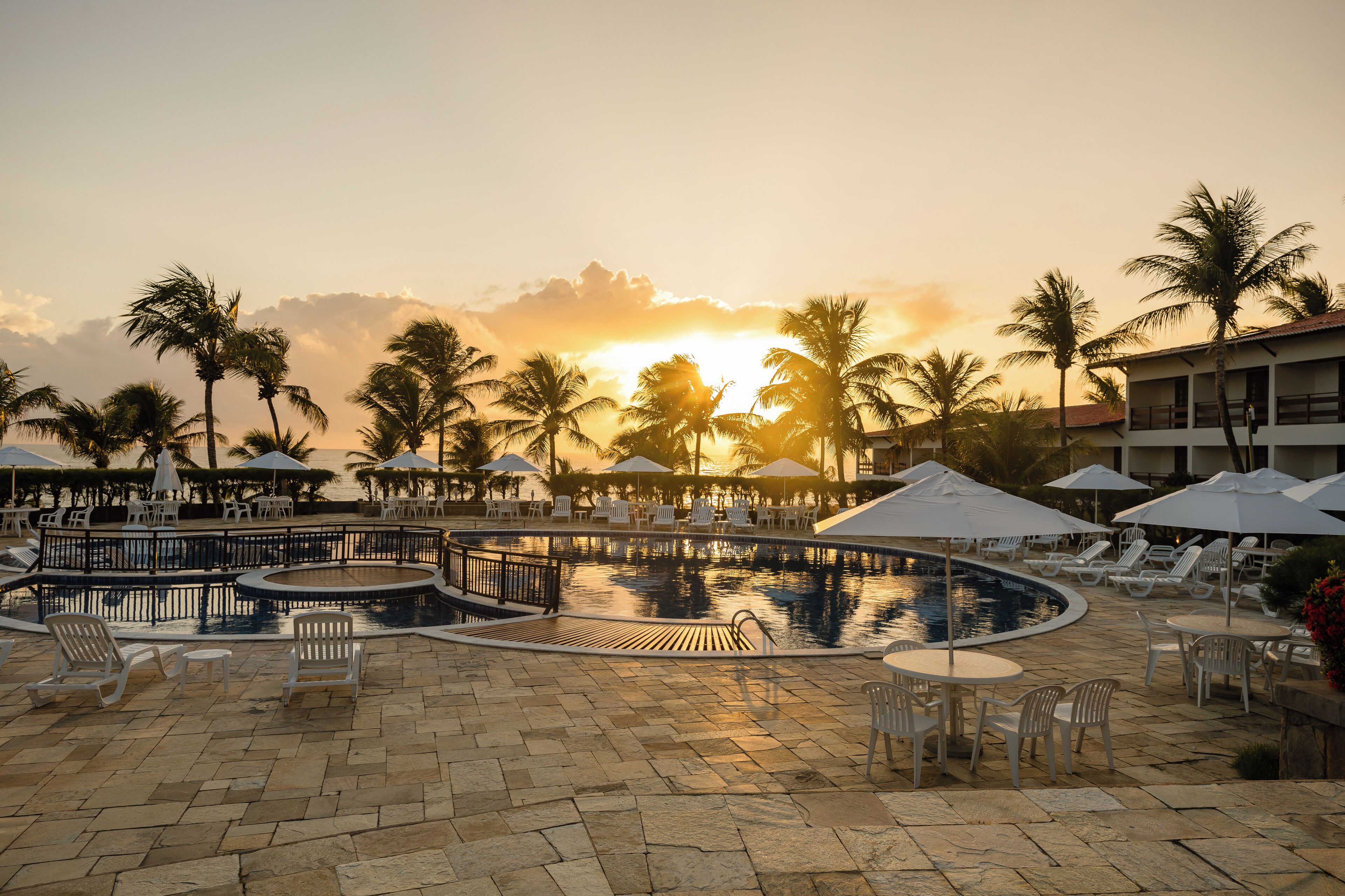 Un área de piscina con sillones de playa y sombrillas, rodeada de palmeras y un edificio al fondo.