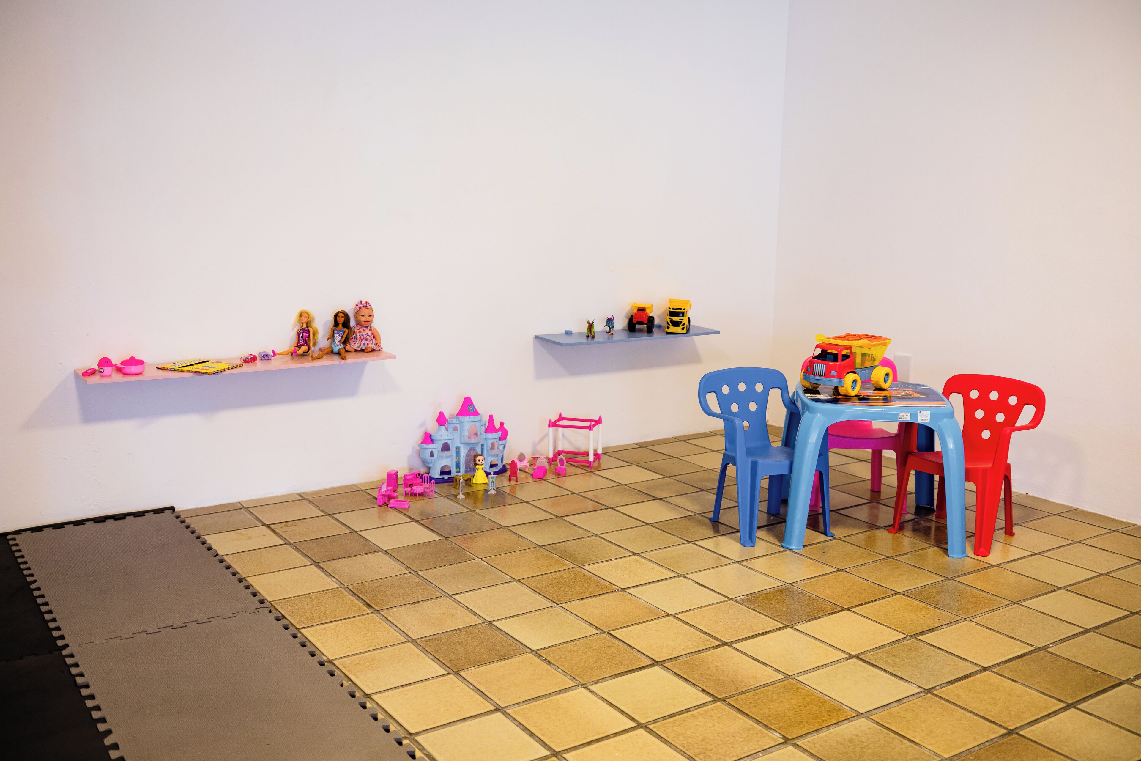 Um espaço de brincadeiras com piso ladrilhado, uma cadeira azul e vermelha, uma mesinha e prateleiras com brinquedos e um modelo de castelo.