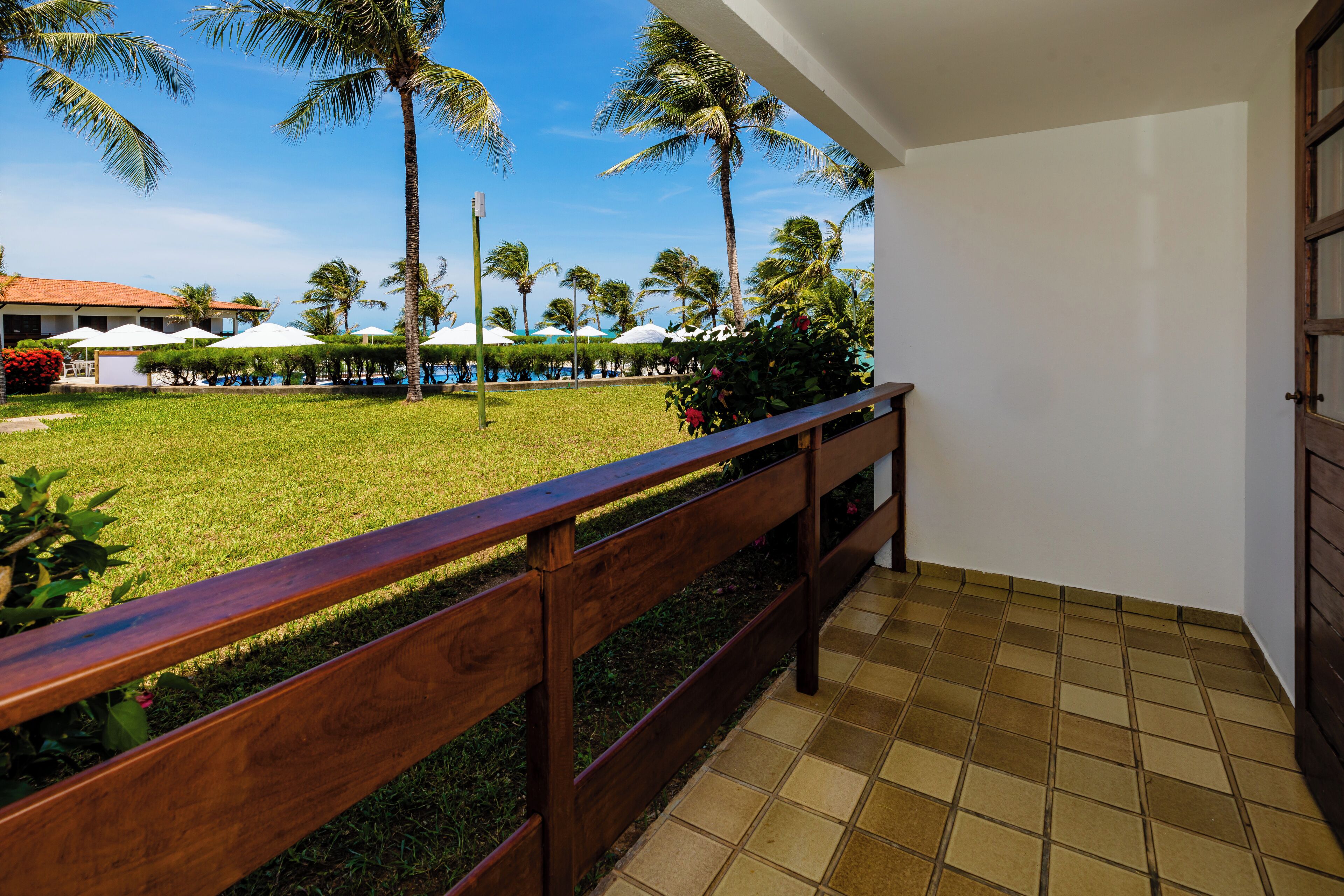 Un balcon doté d’une balustrade en bois donnant sur un espace piscine agrémenté de parasols et de palmiers.