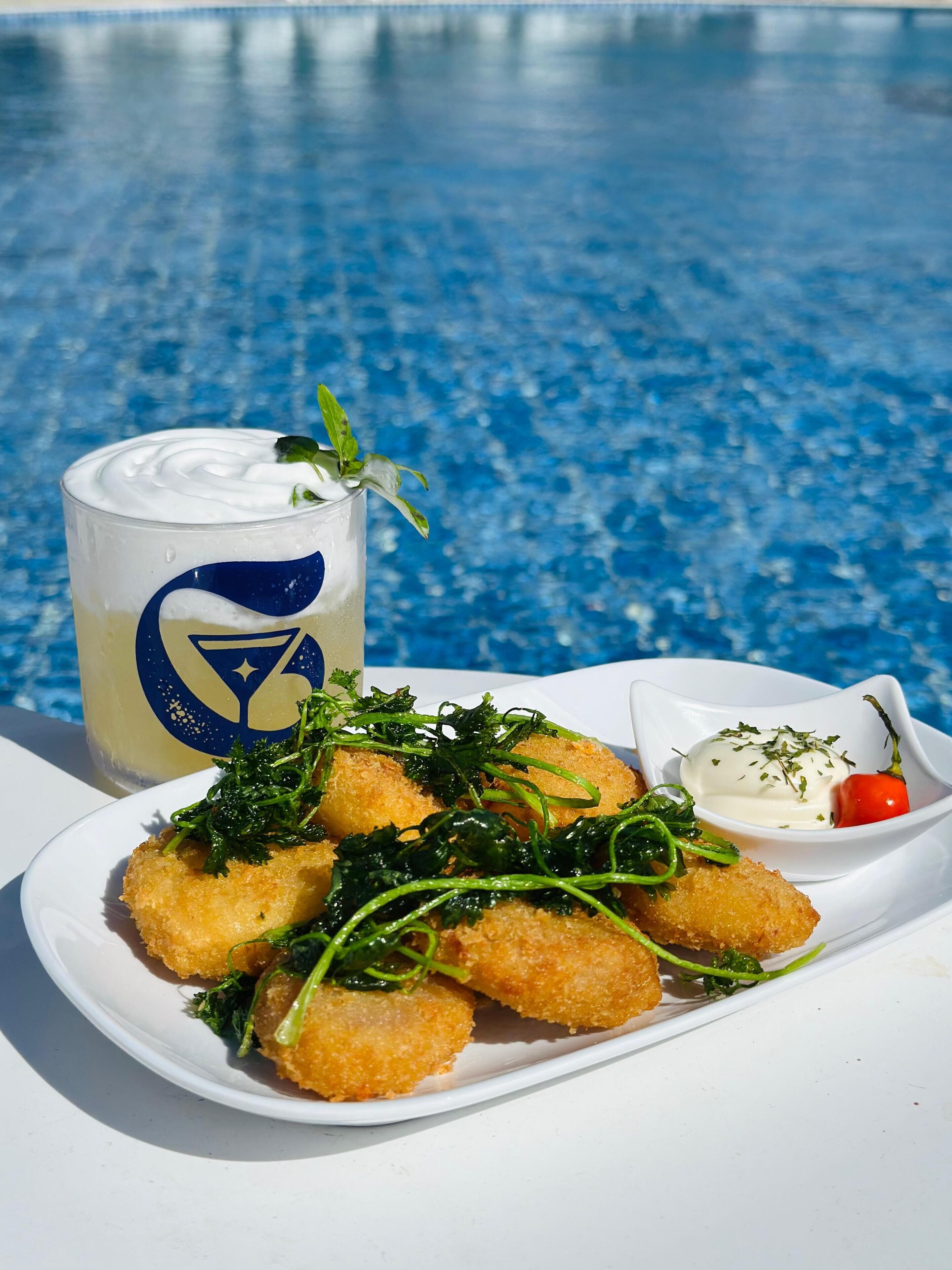 A plate of fried food with greens and a small bowl of sauce, next to a drink with a blue logo.