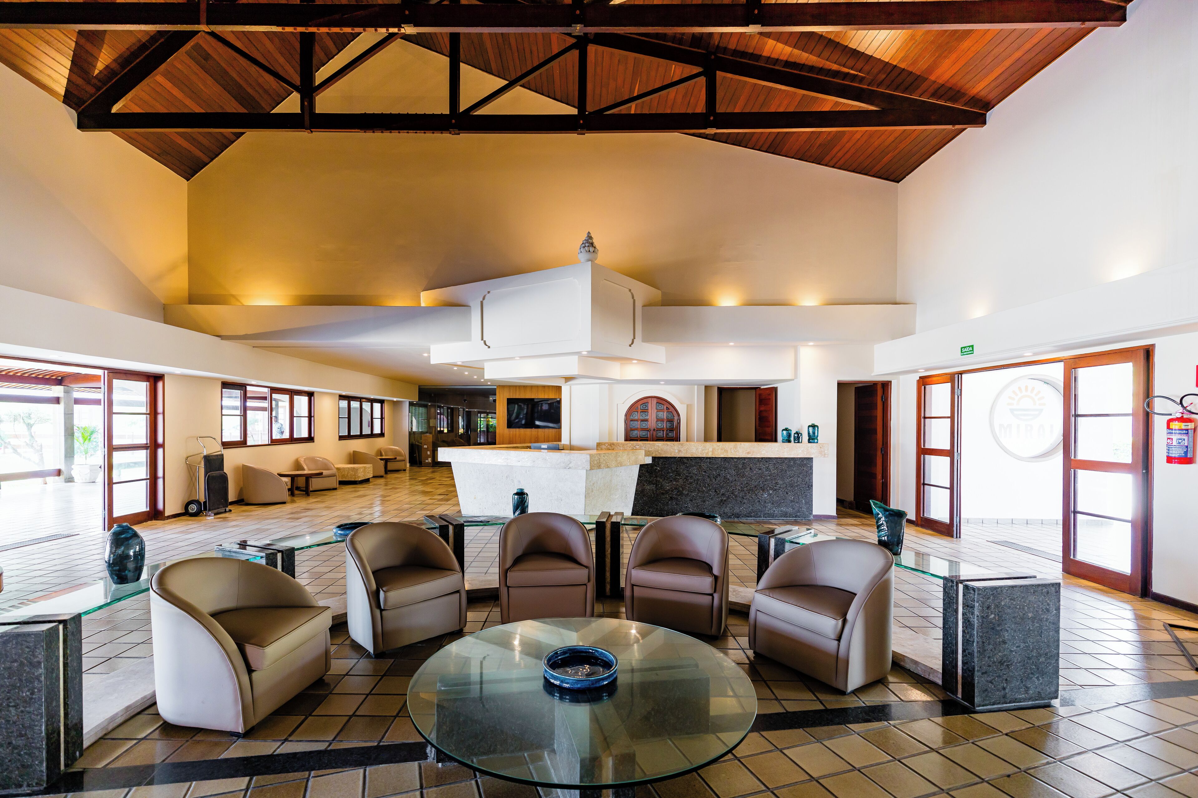 Um hall amplo com teto alto, balcão de recepção, assentos confortáveis e uma grande mesa de vidro.