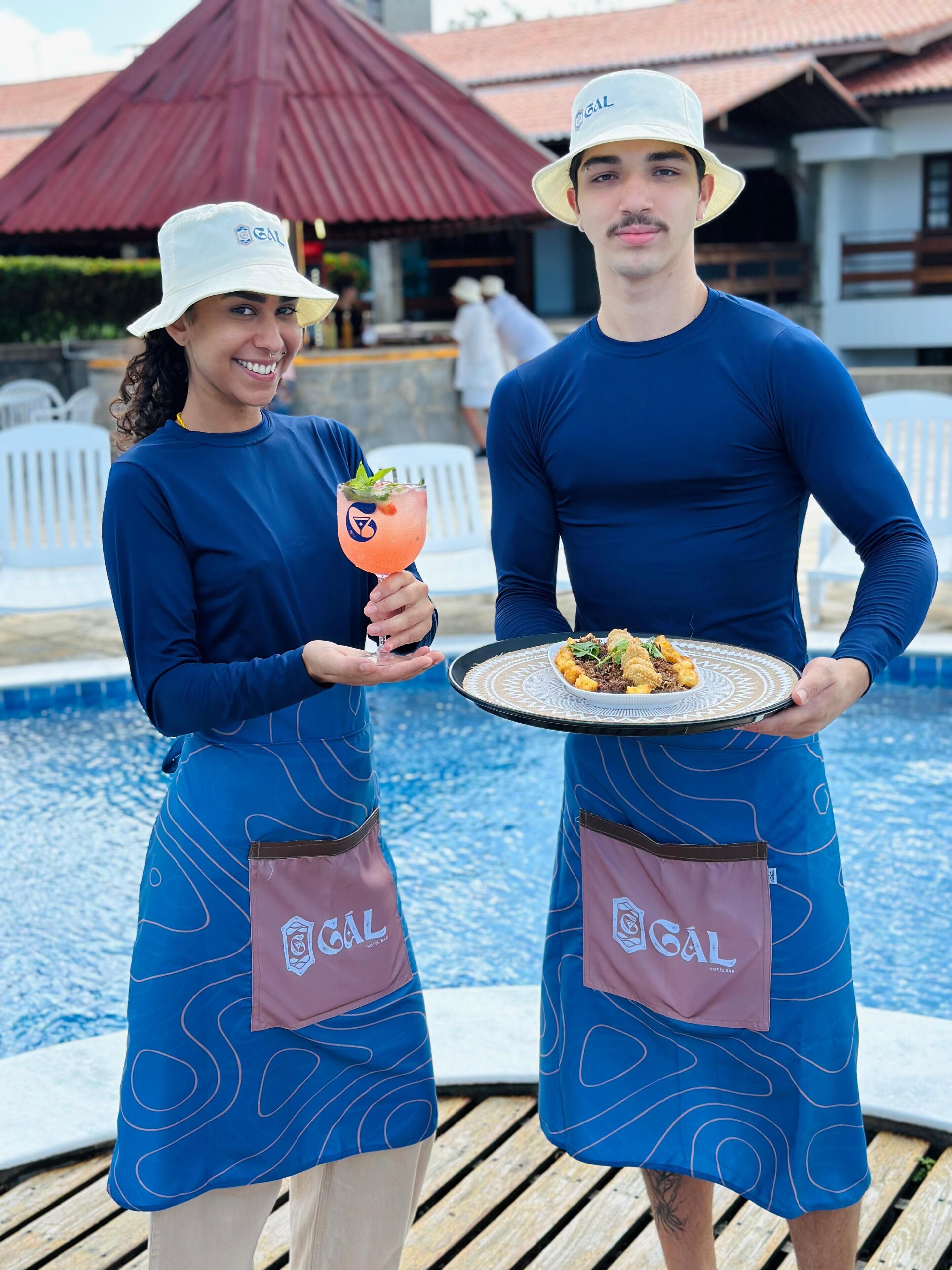 Dois funcionários com uniforme azul e chapéus segurando uma bebida e um prato de comida perto de uma piscina.