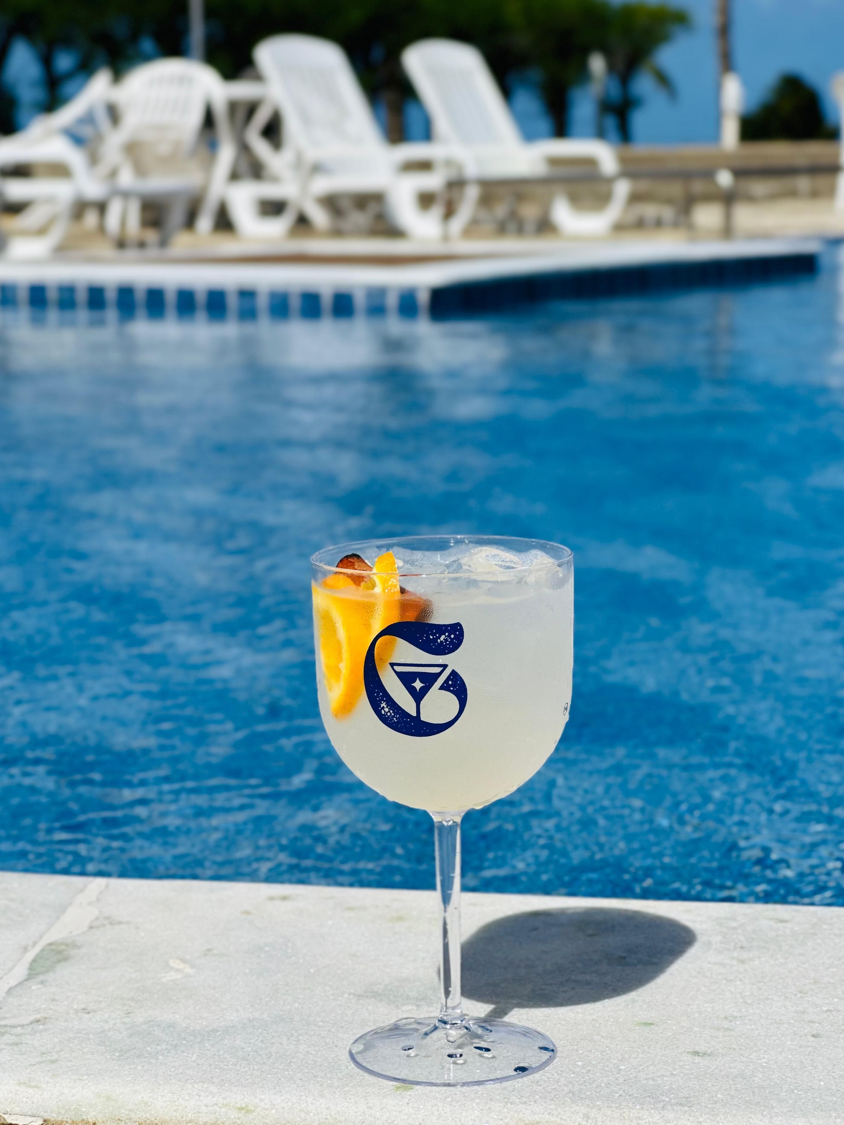 A clear glass with a cocktail garnish and a logo, placed on a poolside with lounge chairs in the background.
