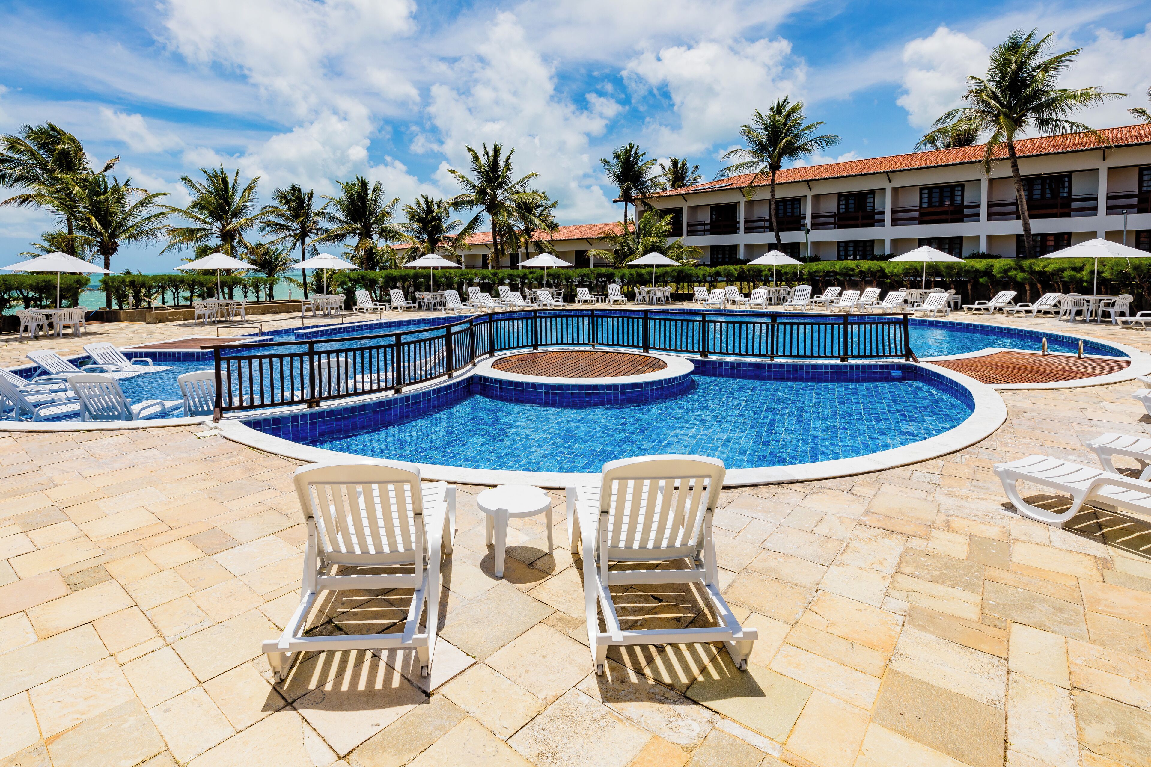 A pool area with lounge chairs, a small table, and a building with a red roof in the background.