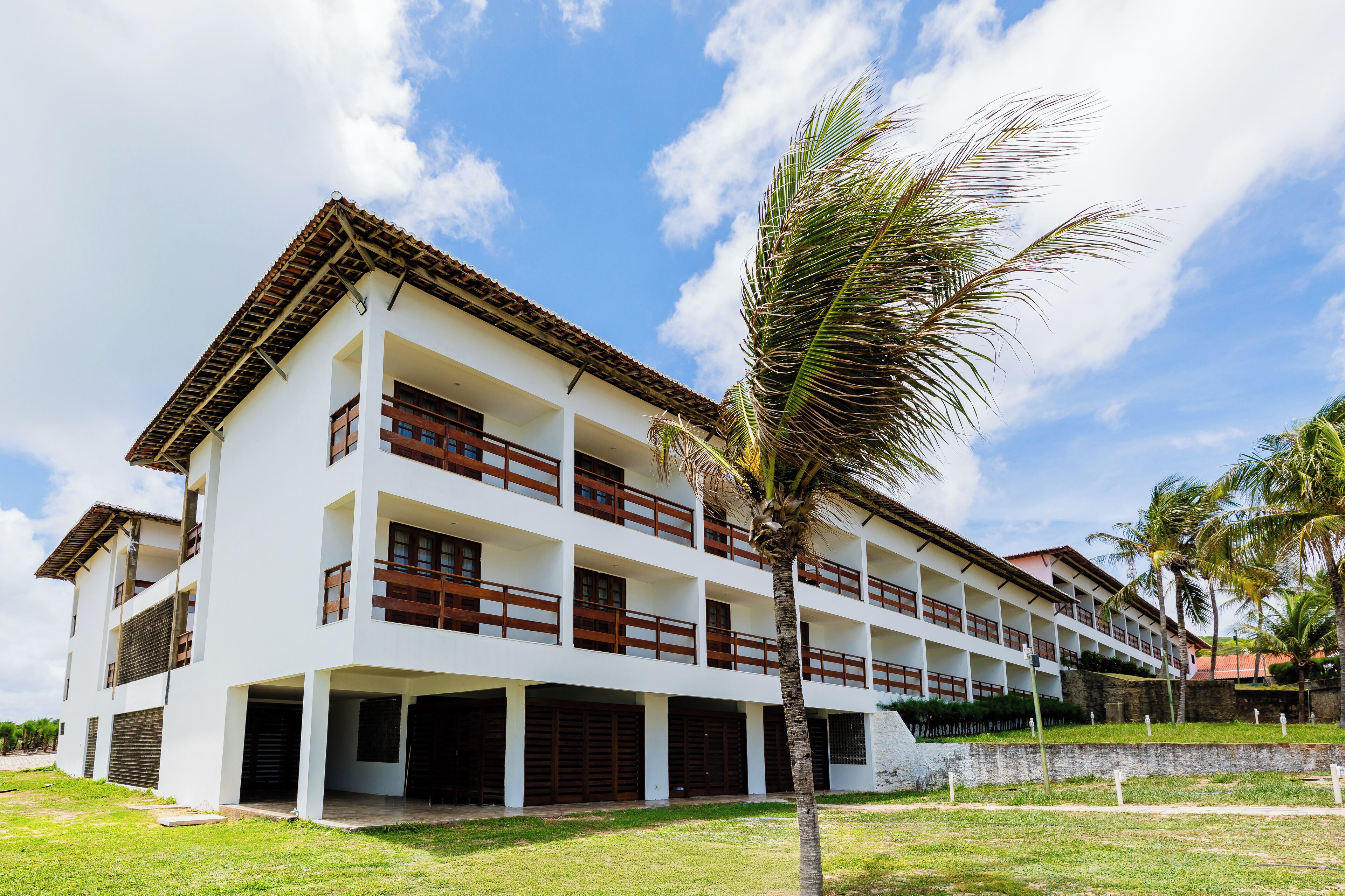 Un hotel de varios pisos con fachada blanca, balcones de madera y una palmera en primer plano.