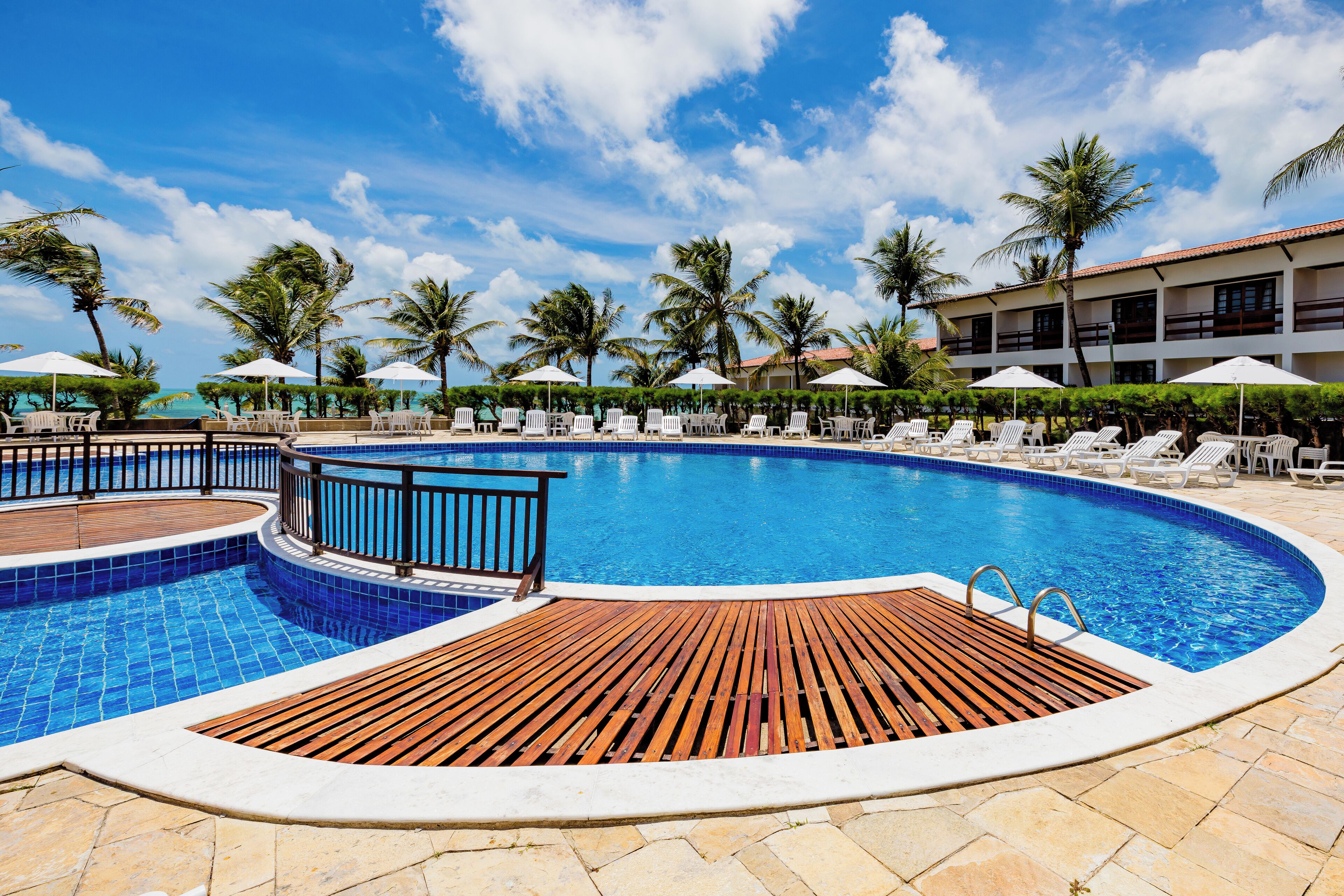A swimming pool with lounge chairs and umbrellas, surrounded by palm trees and a building with multiple balconies.