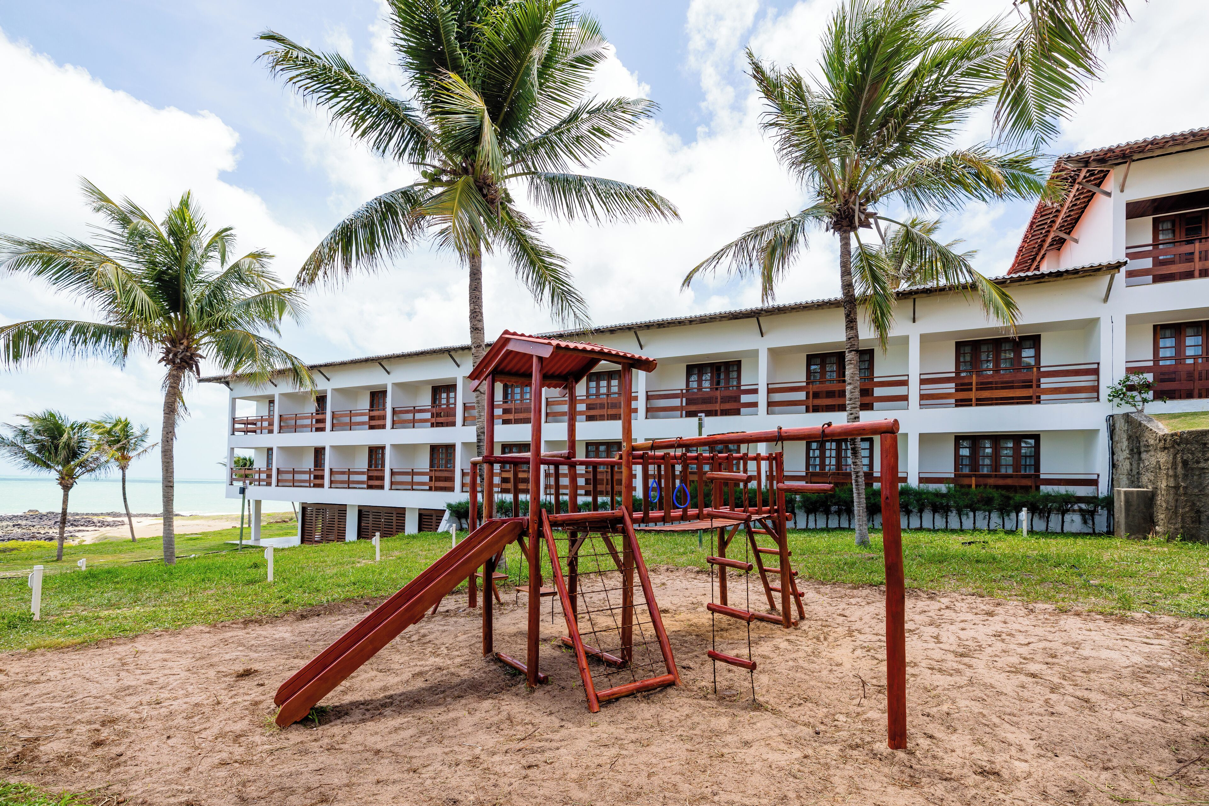 A playground with a slide and climbing structures, surrounded by palm trees and a building with balconies.