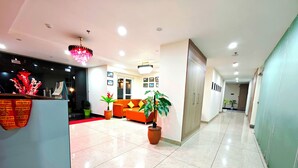 Zona de recepción de hotel con alfombra roja, mostrador de recepción, área de descanso con sofá naranja y plantas en macetas.