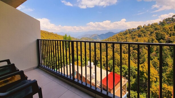 Un balcón con baranda metálica, una banca y vista a un paisaje verde con árboles y montañas a lo lejos.