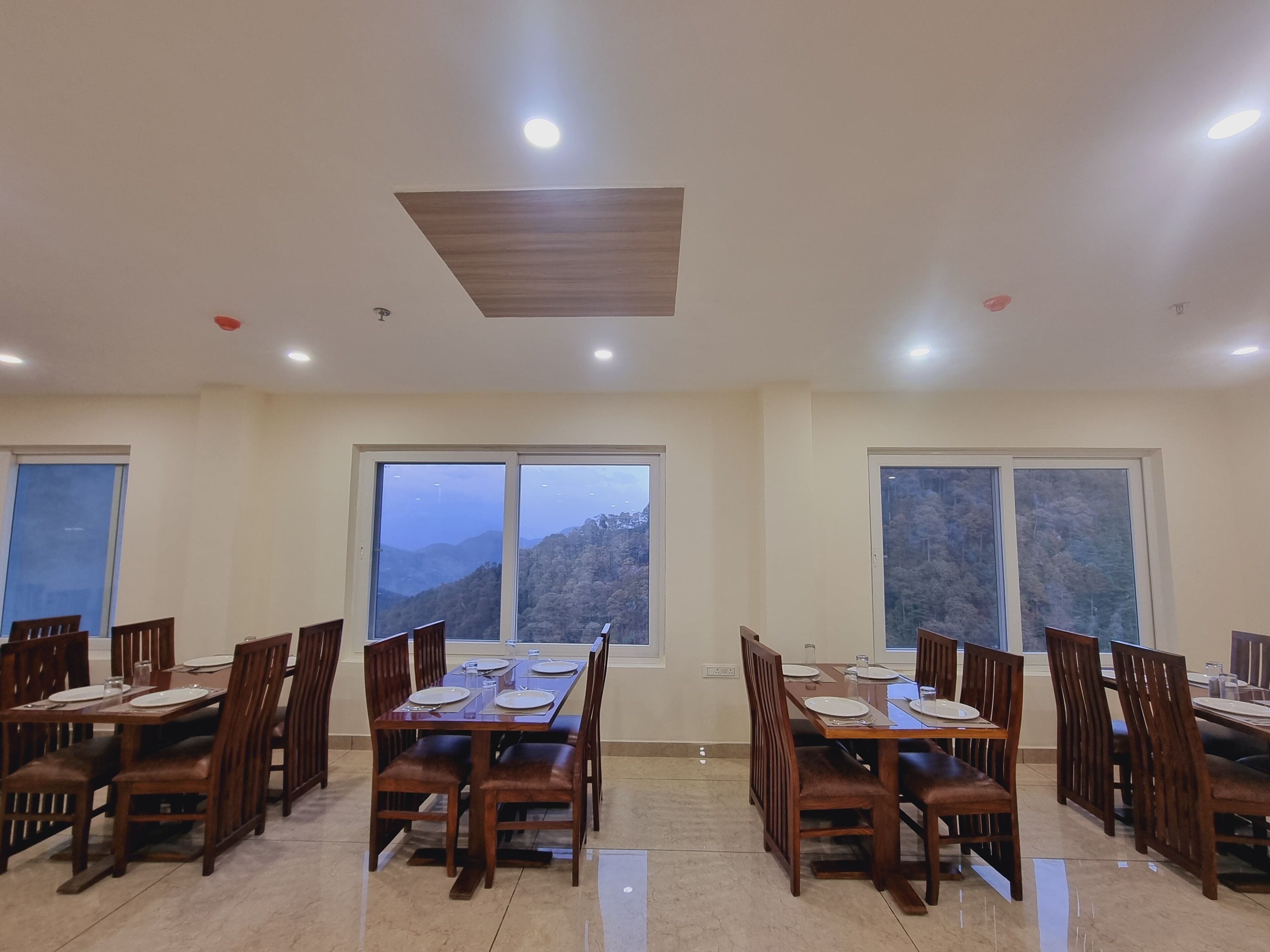 Un comedor con mesas y sillas de madera, amplias ventanas con vistas a las montañas y luces empotradas en el techo.