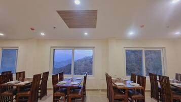 Un comedor con mesas y sillas de madera, amplias ventanas con vistas a las montañas y luces empotradas en el techo.