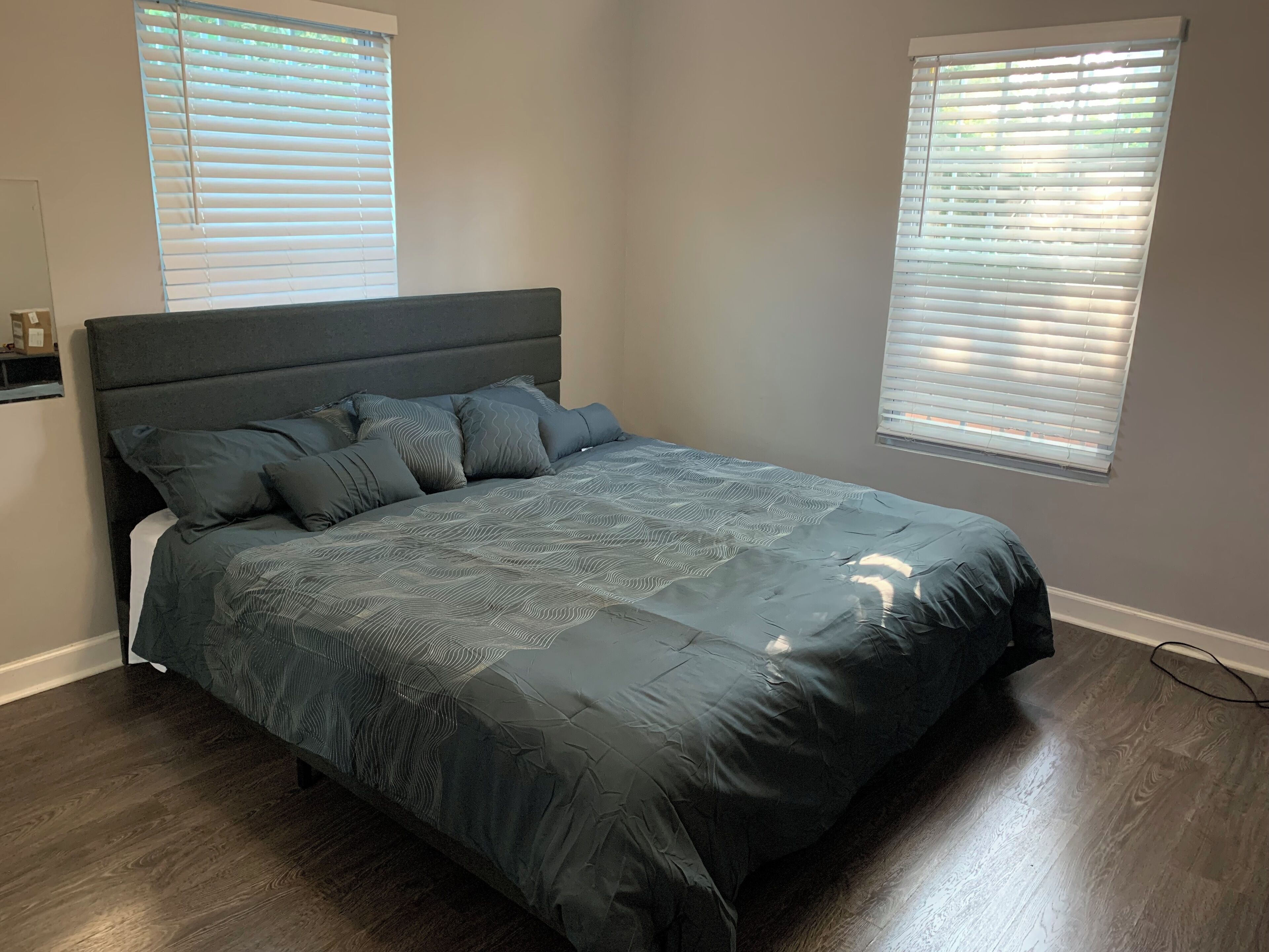 A bedroom with a large bed, two windows with blinds, and a wooden floor.