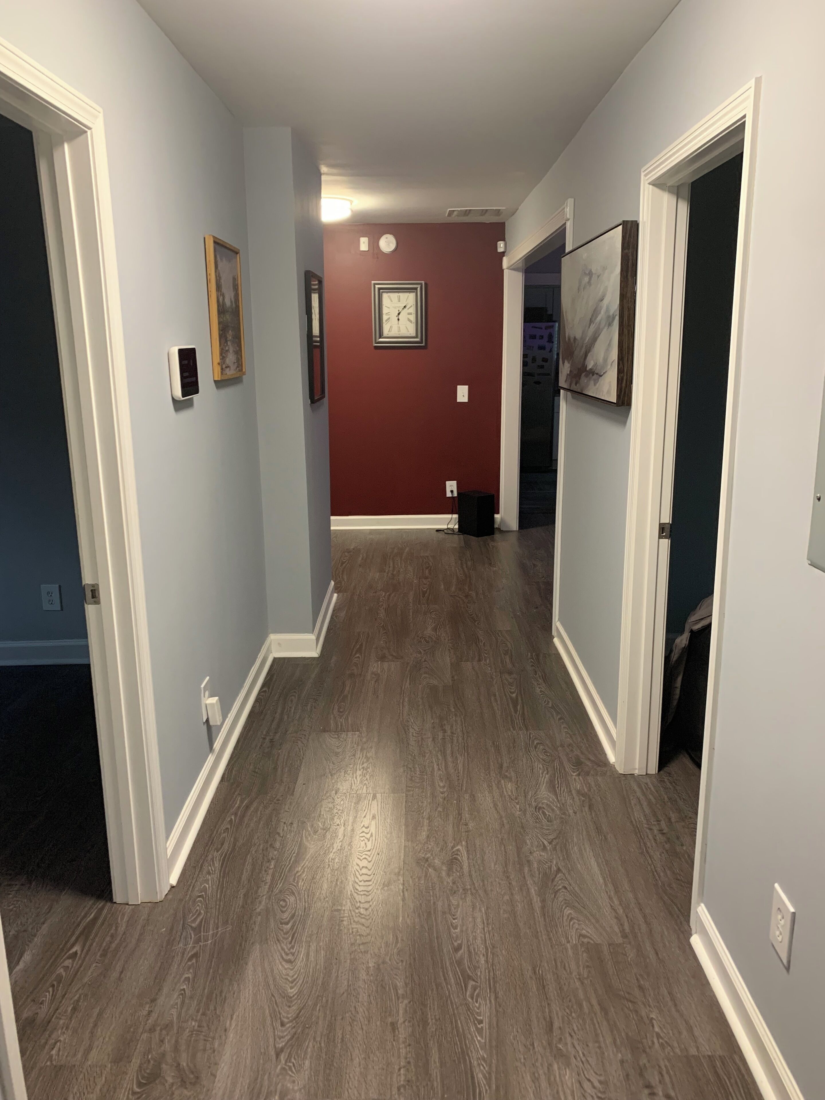 A hallway with a wooden floor, light blue walls, and a red accent wall. There are framed pictures and a clock on the walls.