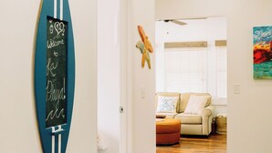 A hallway with a surfboard sign, a ceiling fan, and a couch in the background. - “La Playa” Downtown Brunswick Beach Cottage - near St. Simons & Jekyll  (Brunswick)