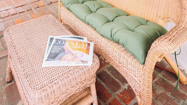 A wicker sofa with a green cushion and a magazine on a matching ottoman, a brick floor, and a window with blinds. - “La Playa” Downtown Brunswick Beach Cottage - near St. Simons & Jekyll  (Brunswick)