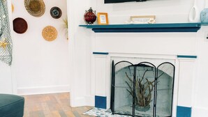 A living room with a flat-screen TV mounted above a fireplace, a blue mantelpiece, and a patterned rug. - “La Playa” Downtown Brunswick Beach Cottage - near St. Simons & Jekyll  (Brunswick)