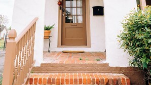 A white building with a brown door, a small porch with steps, and a hanging plant above the door. - “La Playa” Downtown Brunswick Beach Cottage - near St. Simons & Jekyll  (Brunswick)