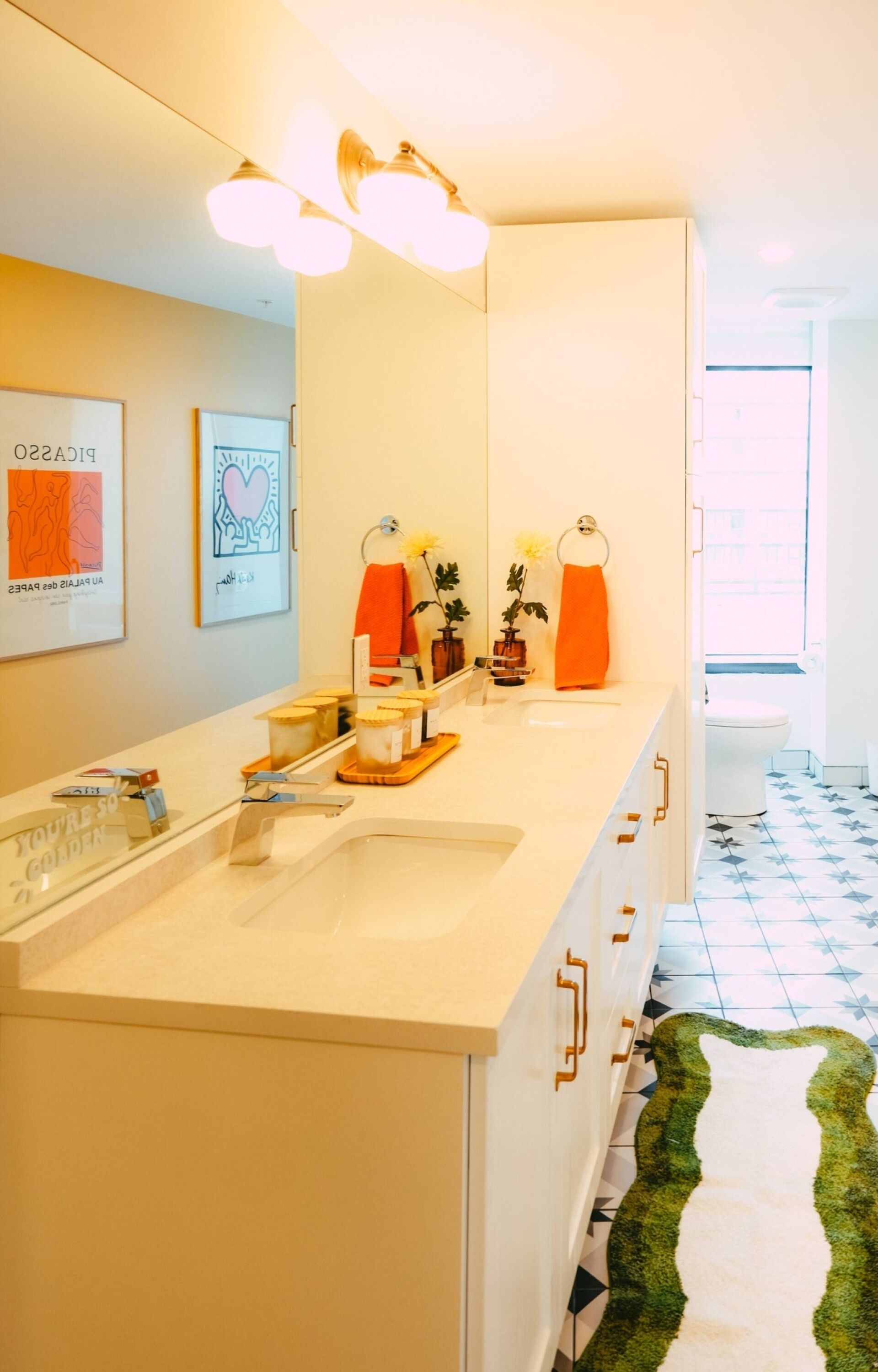 A modern bathroom with a large mirror, a sink, and a toilet. There are framed pictures on the wall and a small plant on the counter.