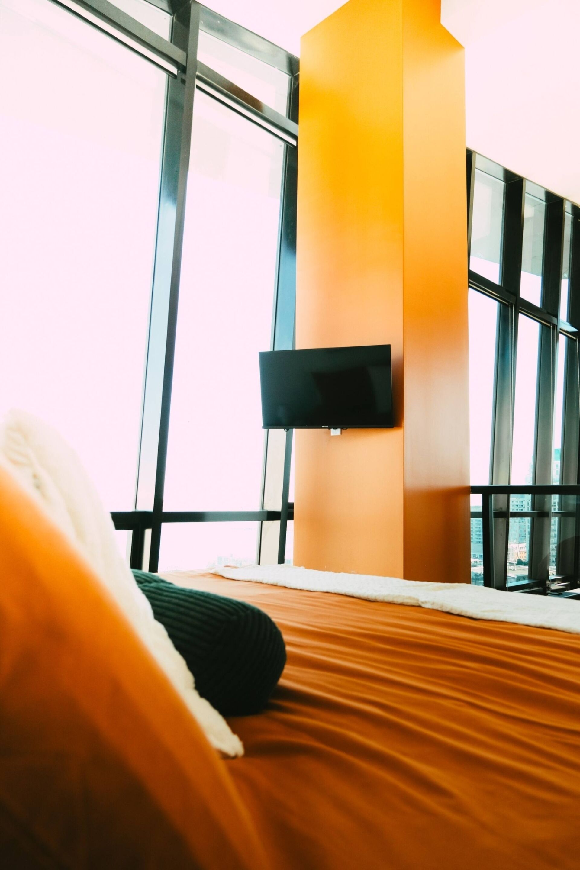 A hotel room with a large window, a flat-screen TV mounted on the wall, and a bed with a white and green pillow.