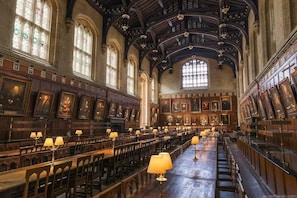 A large hall with rows of wooden benches and tables, high ceilings with intricate woodwork, and large stained glass windows.