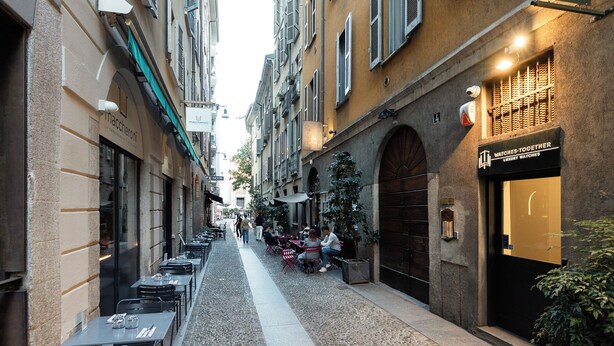 A narrow street with outdoor seating, a watch store, and a restaurant. - Brera Apartments in Arena (Milan)