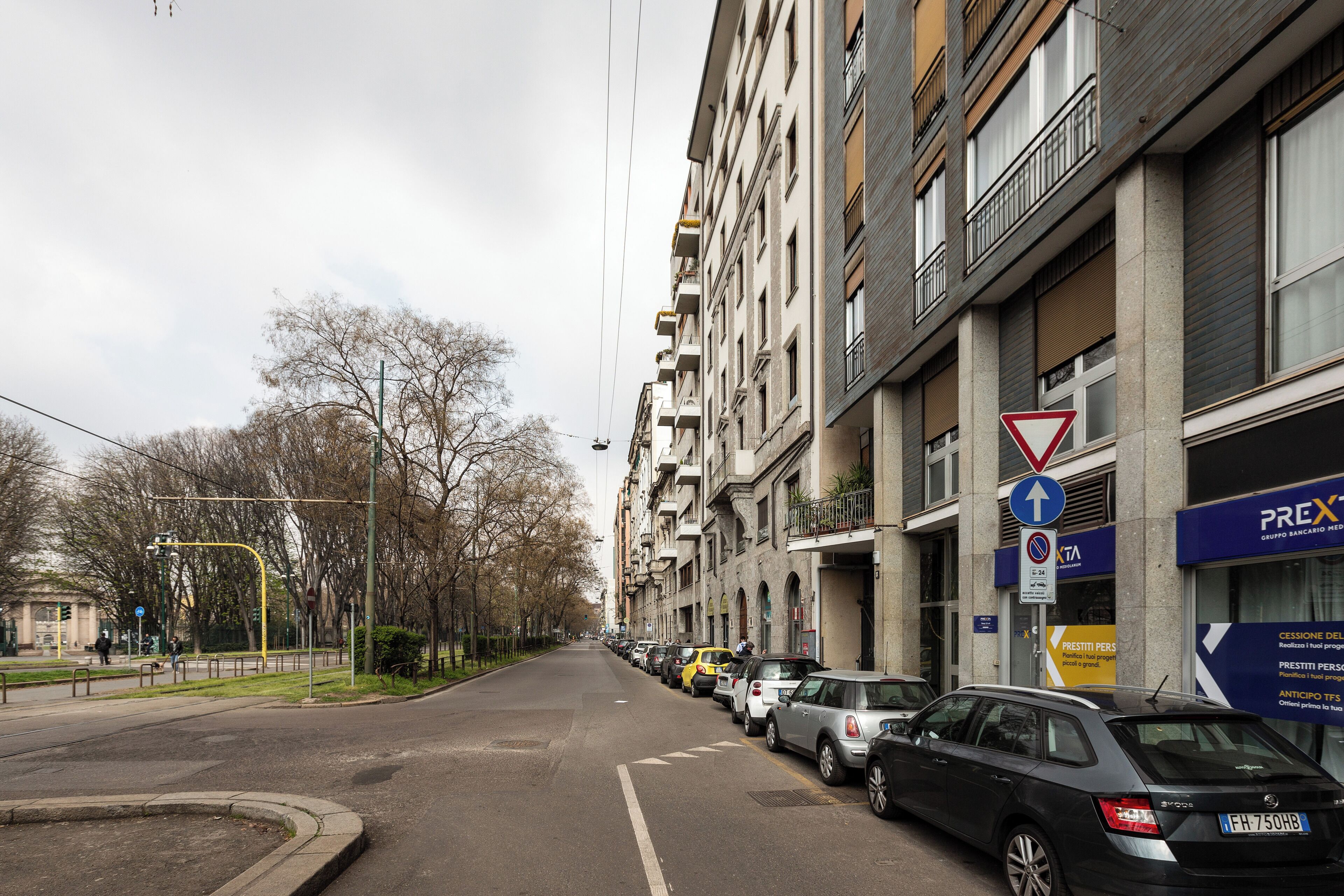 Una strada con auto parcheggiate, un attraversamento pedonale e un edificio con un cartello che indica 'PREX'.