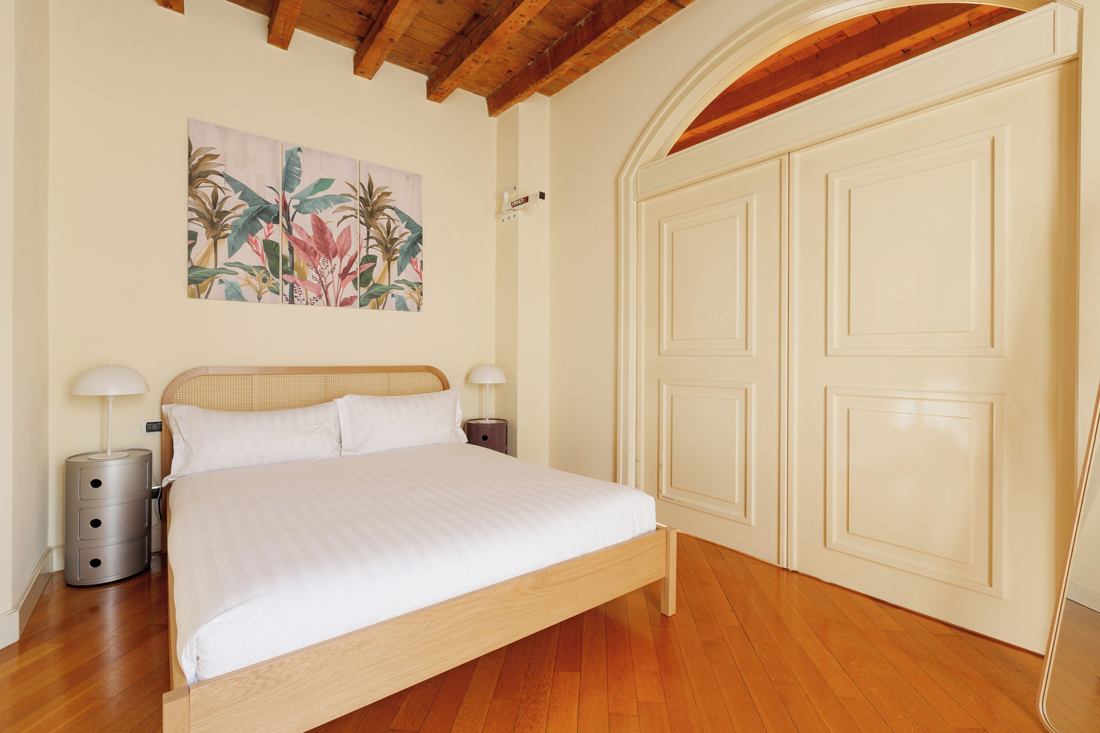 A bedroom with a wooden bed, white walls, a painting of tropical plants, and a white door.