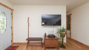 A room with a wooden bench, a small wooden table with a record player, a potted plant, and a flat-screen TV mounted on the wall.