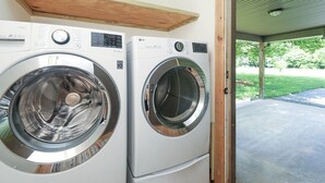 Two front-loading washing machines in a laundry room with a wooden shelf above and a view of a green lawn outside.