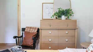 A bedroom with a bed, a wooden dresser with drawers, a chair, a mirror, a framed picture, and a potted plant.