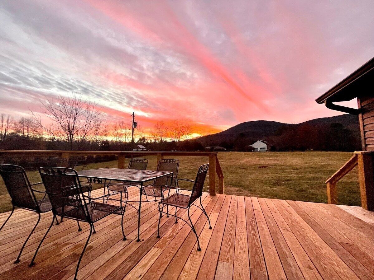 A wooden deck with a table and chairs, overlooking a grassy area and a sunset.