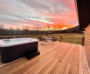 A wooden deck with a hot tub and outdoor furniture, overlooking a grassy area and a sunset.