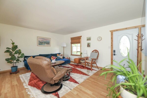 A living room with a brown leather chair, a blue sofa, a wooden coffee table, and a red and white rug. There are two potted plants, a wall clock, and a door with a wooden frame.
