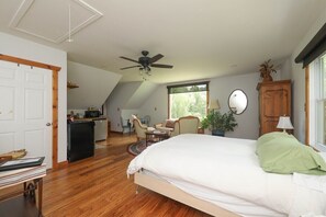 A bedroom with a large bed, a ceiling fan, a dining table, and a kitchen in the background.