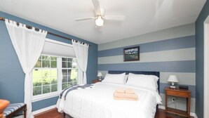 A bedroom with a large bed, a ceiling fan, a nightstand with a lamp, and a window with curtains. - Blue Rivers Cottage and Loft (Afton)