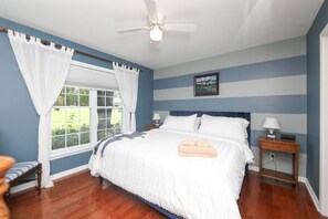 Un dormitorio con una cama grande, un ventilador de techo, una mesita de noche con lámpara y una ventana con cortinas.