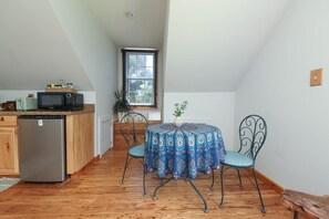 A compact kitchen with a refrigerator, microwave, and wooden cabinets. A dining table with a patterned tablecloth and metal chairs is in the center.
