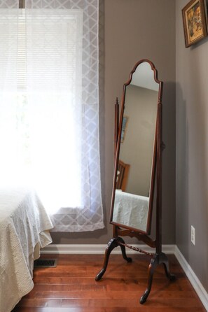 A bedroom with a wooden floor, a bed with a white bedspread, a window with sheer curtains, and a wooden dresser with a mirror. - Blue Rivers Cottage and Loft (Afton)