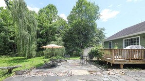 A stone patio with outdoor furniture, a wooden deck, and a green house in the background. - Blue Rivers Cottage and Loft (Afton)