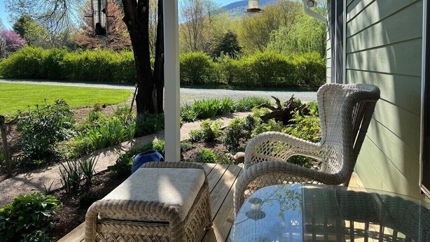A covered porch with wicker furniture and a glass table, overlooking a garden with a bird feeder and a mountain in the background. - Blue Rivers Cottage with Mountain Views (Afton)