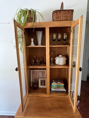 A wooden cabinet with glass doors, containing various items like a vase, wine glasses, a bowl, and books. - Blue Rivers Cottage with Mountain Views (Afton)