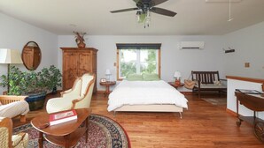 A spacious bedroom with a large bed, a ceiling fan, a wooden wardrobe, a bedside table, a nightstand, a chair, a small table with a book, and a potted plant.