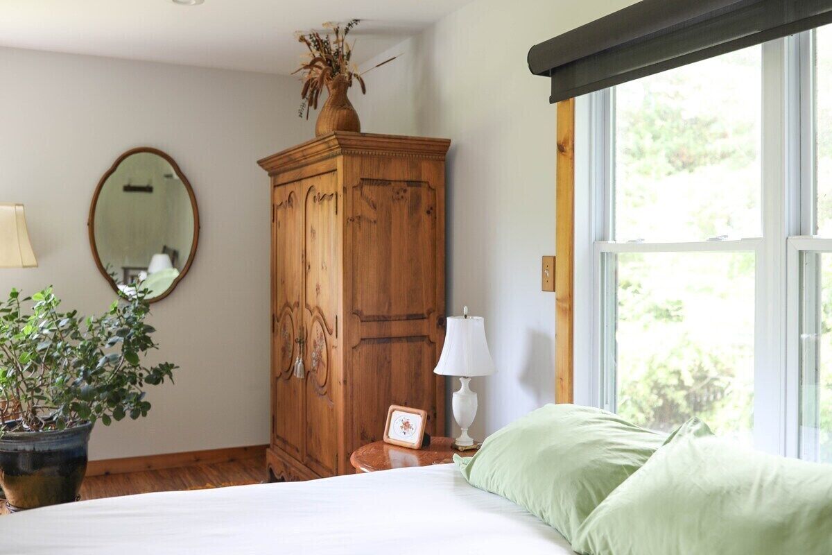 A bedroom with a wooden wardrobe, a bed with green pillows, a bedside table with a lamp, a potted plant, and a mirror on the wall.