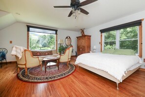 A bedroom with a bed, two chairs, a table, a ceiling fan, and large windows.