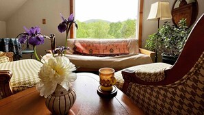 A cozy living room with a sofa, a patterned armchair, a wooden table with a vase of flowers, a lamp, and a view of greenery through a window.