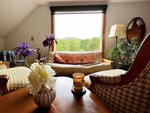 A cozy living room with a sofa, a patterned armchair, a wooden table with a vase of flowers, a lamp, and a view of greenery through a window. - Blue Rivers Loft on the Brew Ridge Trail (Afton)