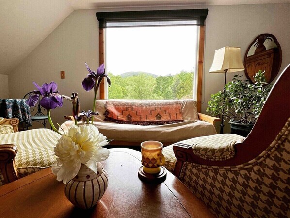 Un salon chaleureux avec un canapé, un fauteuil à motifs, une table en bois ornée d’un vase de fleurs, une lampe et une vue sur la verdure par la fenêtre.