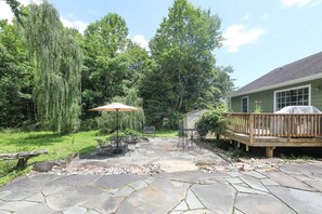 A stone patio with outdoor furniture, a wooden deck, and a green house in the background.