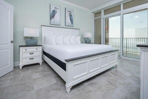 Quarto com cama branca, mesa de cabeceira com abajur e uma janela grande com vista.